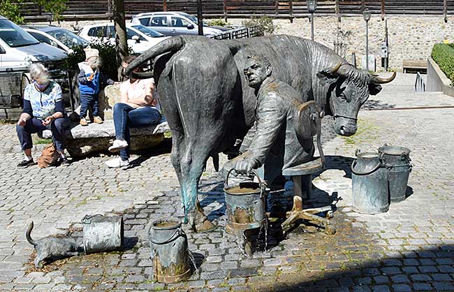 Gebrüder Immler - Steuerzahlerbrunnen Isny Altstadt. Ein Finanzbeamter wundert sich, dass nichts brig bleibt, weil er mit löchrigen Eimern sien Kuh (Steuerzahler) melkt.